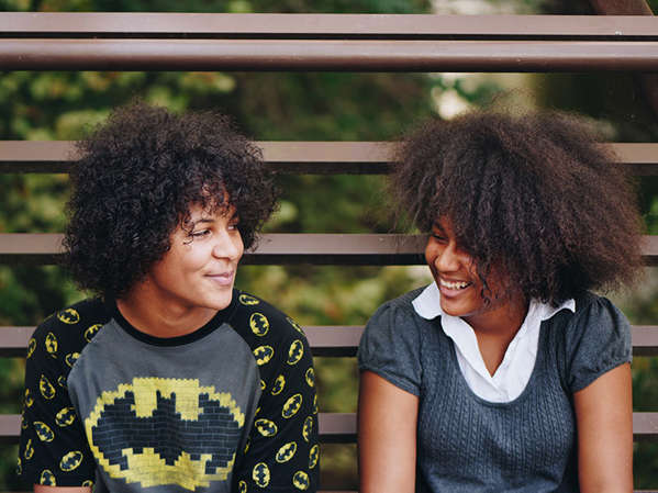Two teenage siblings with dark curly hair sit outside on bleachers looking at one another smiling. 