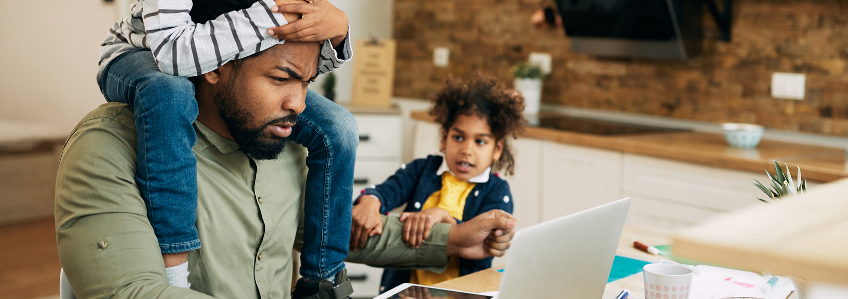 stressed Black foster dad searches for help on his laptop while kids tug on his arm